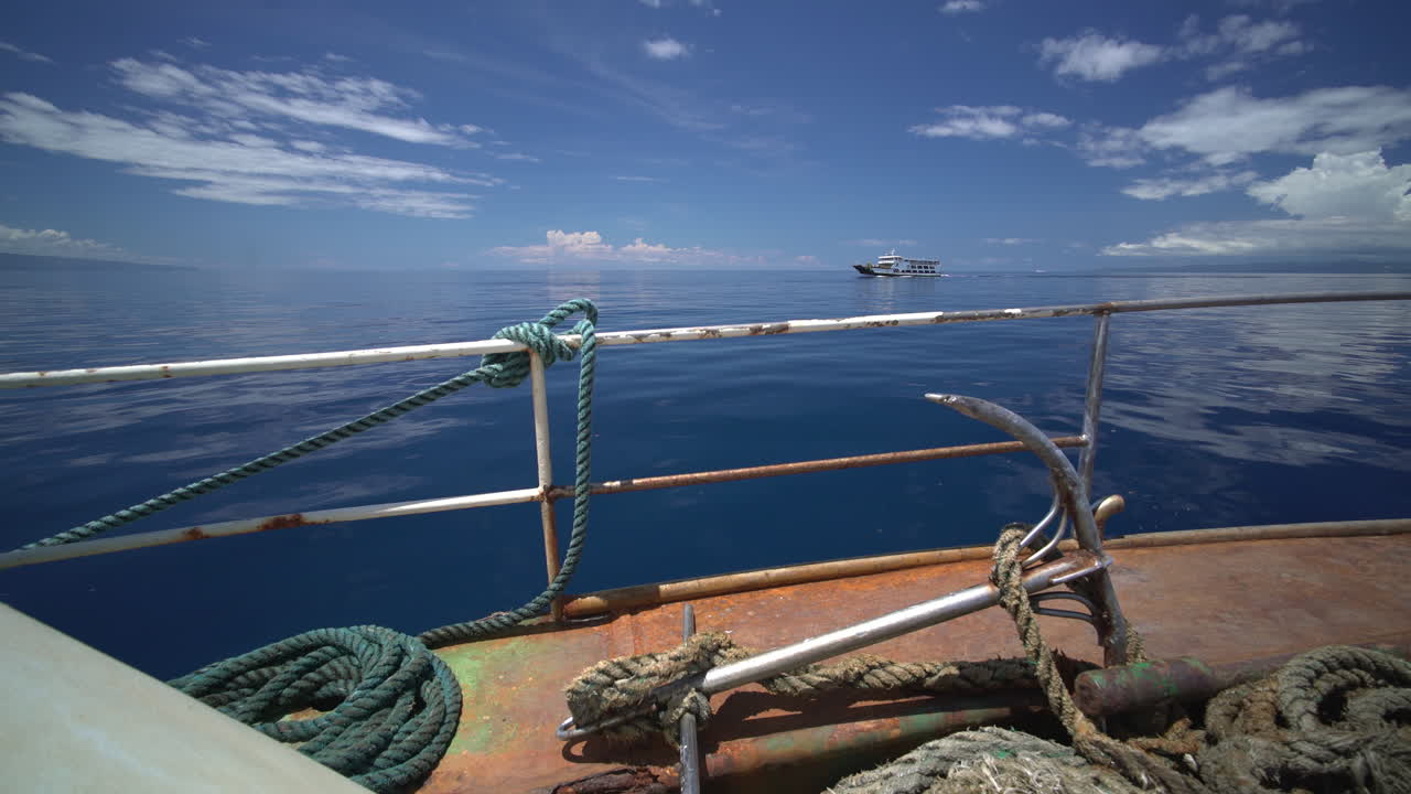 Blick vom Bootsdeck mit Seilen und Anker während der Fahrt über den ruhigen Pazifik, ein weiteres Boot fährt am fernen Horizont unter klarem Himmel vorbei.