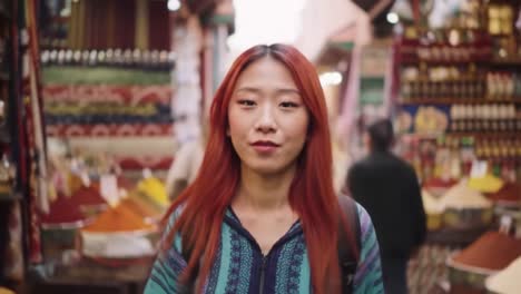 a young woman walks through colorful market stalls filled with spices and local goods. the atmosphere is lively, with vendors and shoppers all around, showcasing cultural richness.