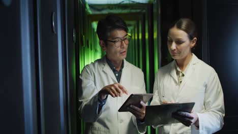 diverse female and male it technicians in lab coats using tablet and laptop checking computer server