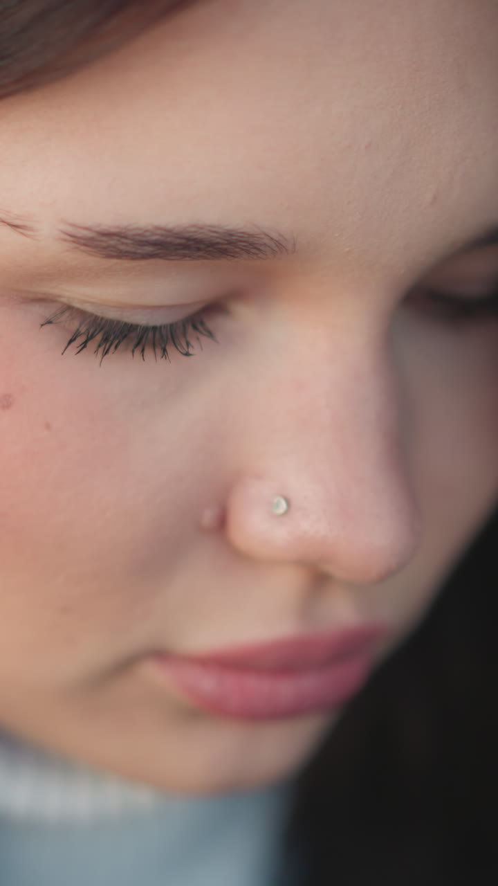 primer plano de una mujer blanca chic con labios rosados, centrándose en algo delante de ella, con un anillo sutil en la nariz y un pendiente de aro dorado, ojos bajados, expresión suave