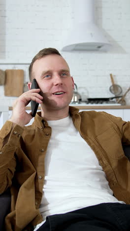 Young man talking the phone in the kitchen. Guy is sitting on sofa with white mug and speaking on a mobile phone at home. Vertical video