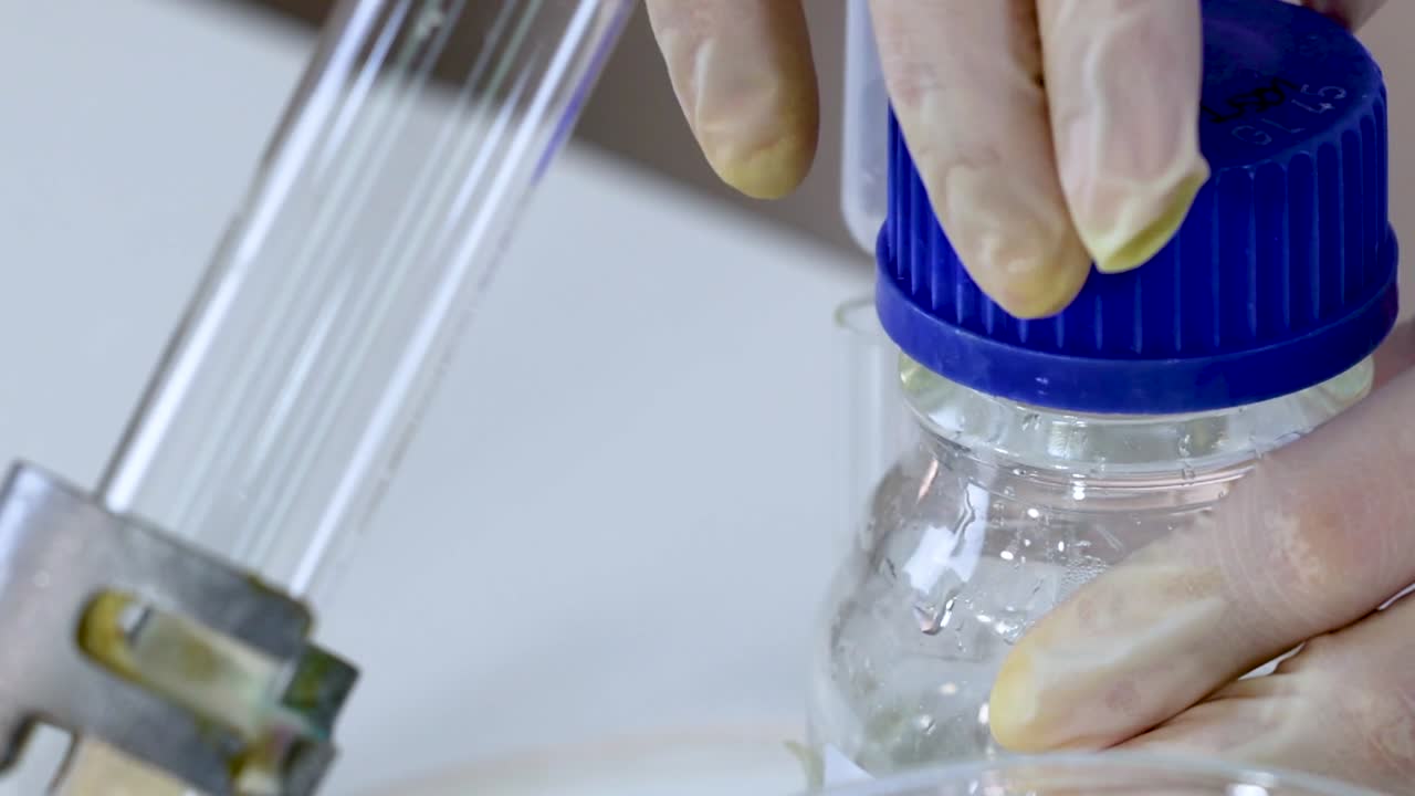 Close-up of gloved hands handling a jar with a blue cap and a glass tube in a laboratory setting.