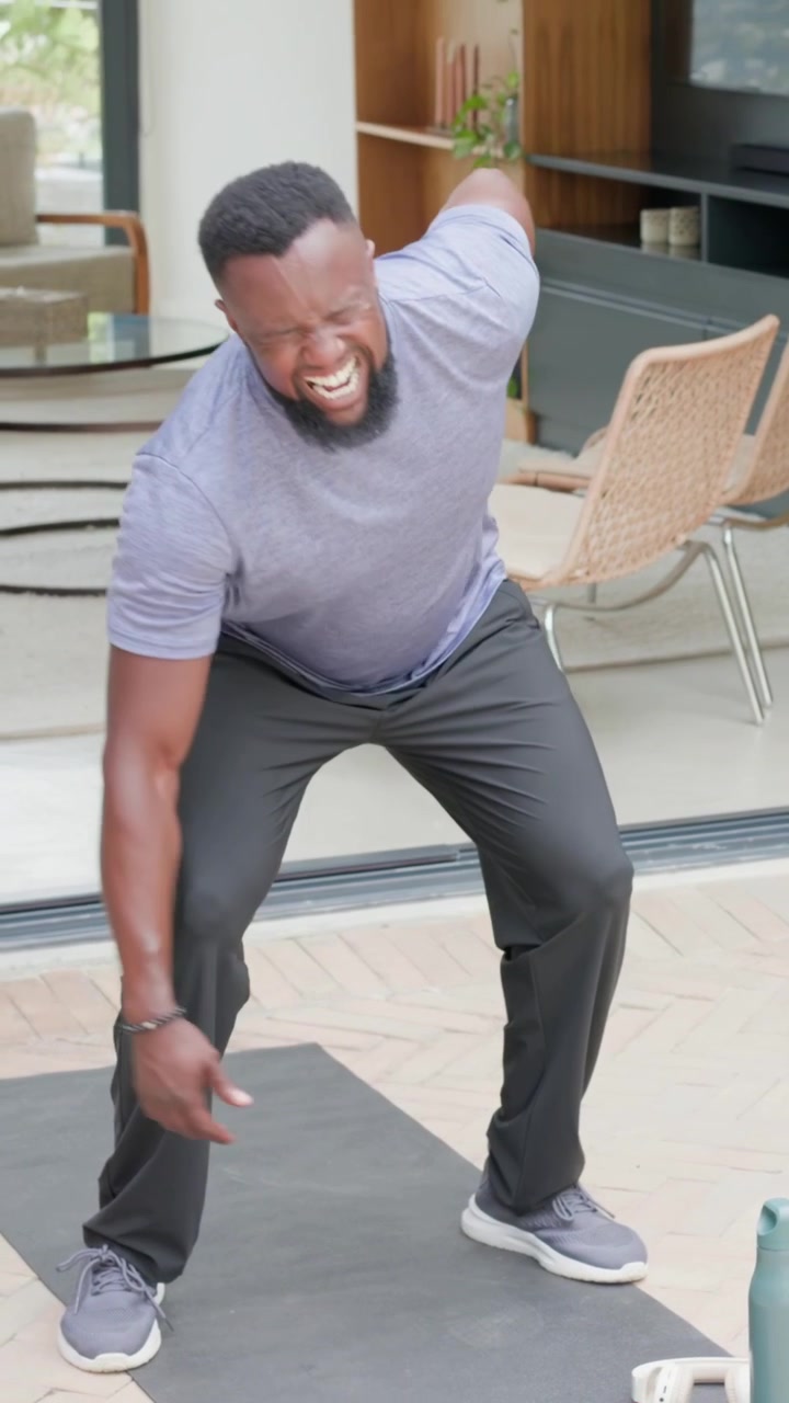 Vertical video: Squatting man in gray tee reaching white weight on home mat, easing back pain