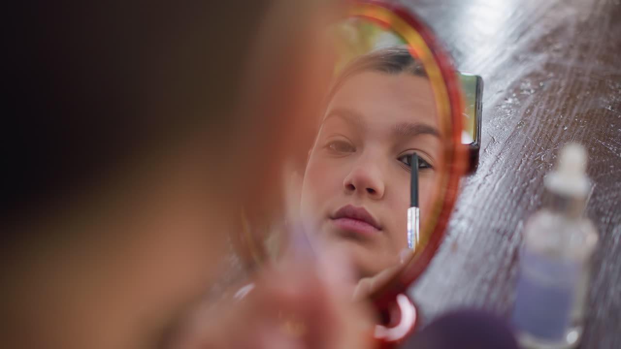 Mirror reflection of young lady applying eye pencil to her eye, focus on precise makeup application with blurred background, highlighting personal beauty routine in a casual setting