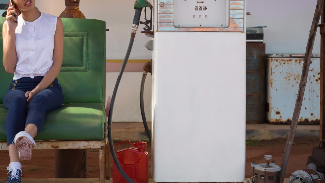 mujer biracial hablando en un teléfono inteligente sentada junto a una bomba de combustible en una gasolinera