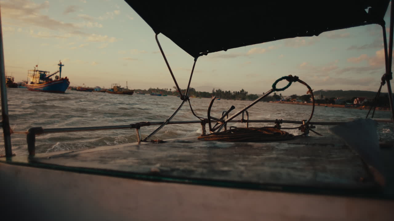 Ponte di una piccola barca con cime e tendalino che si muove su un mare calmo vicino a un villaggio costiero al tramonto.