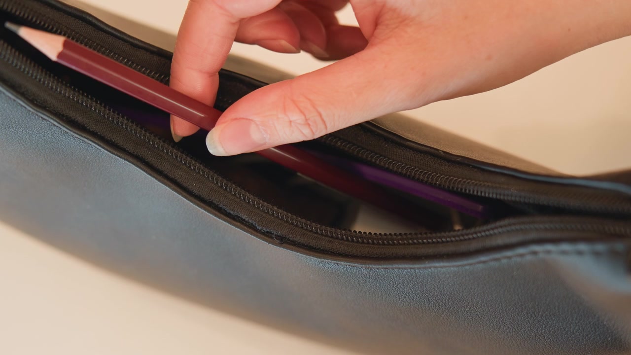 Una mano en primer plano coloca una brocha de maquillaje con mango de madera en un estuche de cuero oscuro con cremallera y forro morado, mostrando un almacenamiento cuidadoso, apertura parcial, dedos en el mango, luz cálida y material táctil.