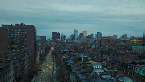 Forwards-fly-above-rows-of-red-brick-apartment-houses-in-residential-urban-neighbourhood-at-twilight.-High-rise-downtown-buildings-in-background.-Boston,-USA