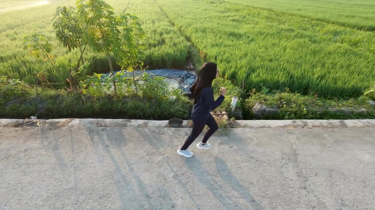 High angle side profile of a Muslim woman wearing a hijab jogging on a rural concrete path next to lush green rice fields. Healthy outdoor fitness and active lifestyle in the morning sunlight.