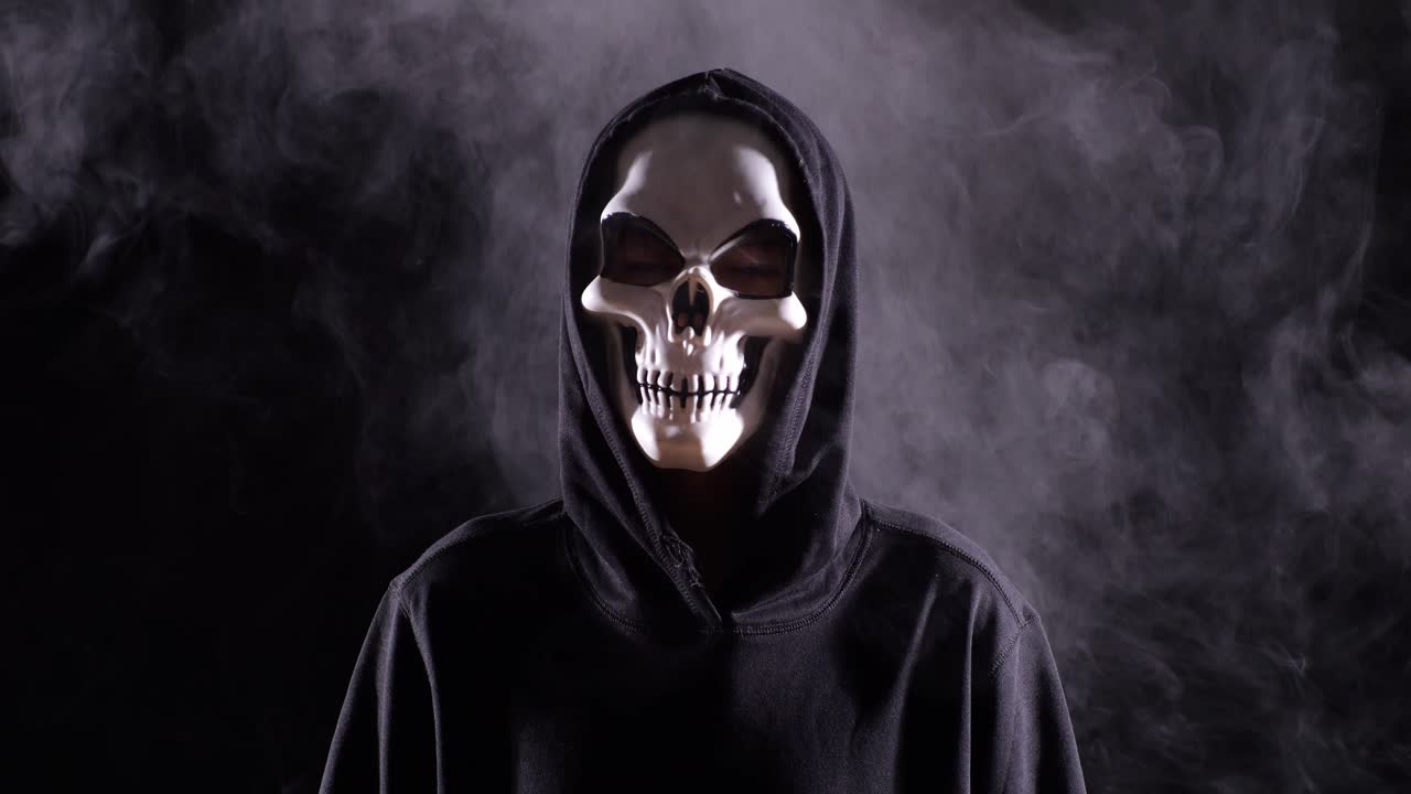 cerca de hombre aterrador en la sudadera con capucha usando máscara de halloween de pie en el fondo negro con humo