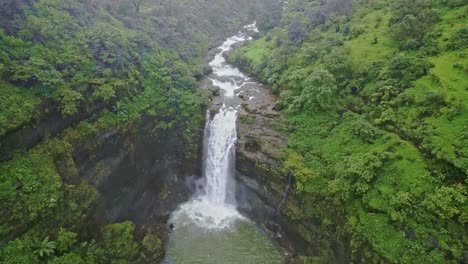 a breathtaking drone view of a majestic waterfall cascading into a serene river, surrounded by lush green mountains and dense forests with the misty monsoon sky in bhandardara maharashtra india