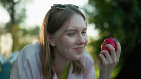 close up of young woman with glasses resting on her head holding an eaten red apple, chewing softly with a warm smile and relaxed demeanor, surrounded by greenery and soft bokeh light