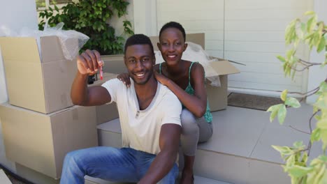 Portrait-of-smiling-african-american-couple-holding-keys-sitting-outside-house-with-packing-boxes