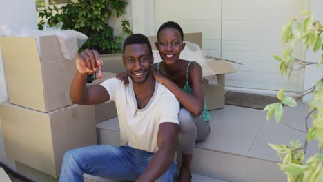 portrait of smiling african american couple holding keys sitting outside house with packing boxes