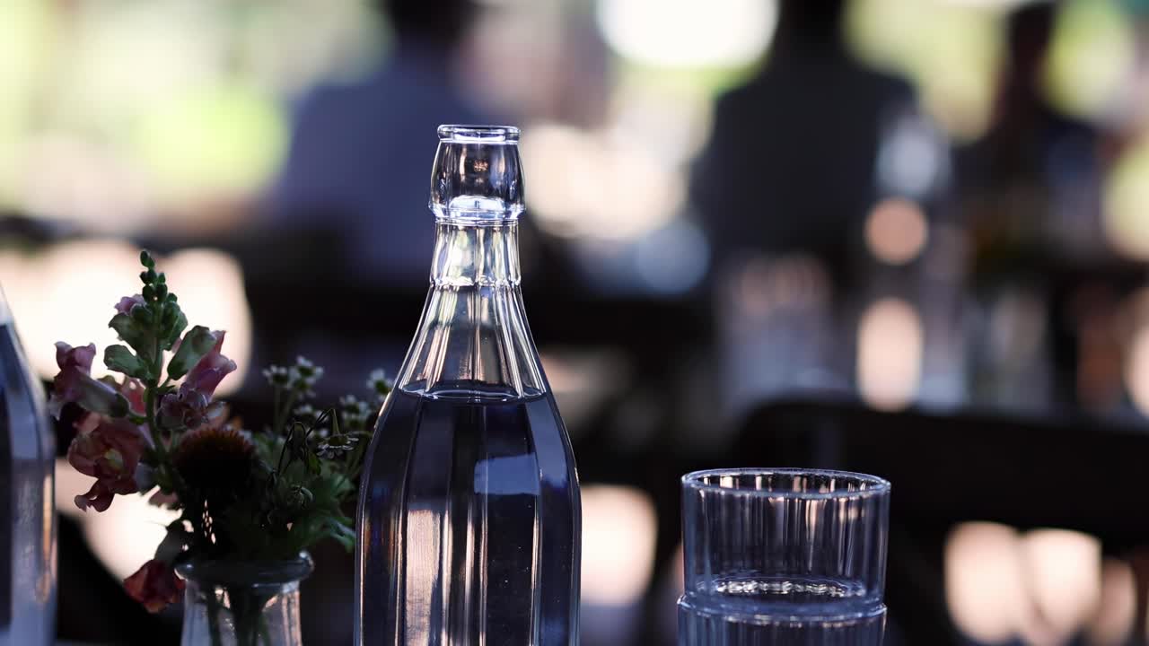 A clear glass bottle and a floral arrangement sit elegantly on a table with a blurred background.