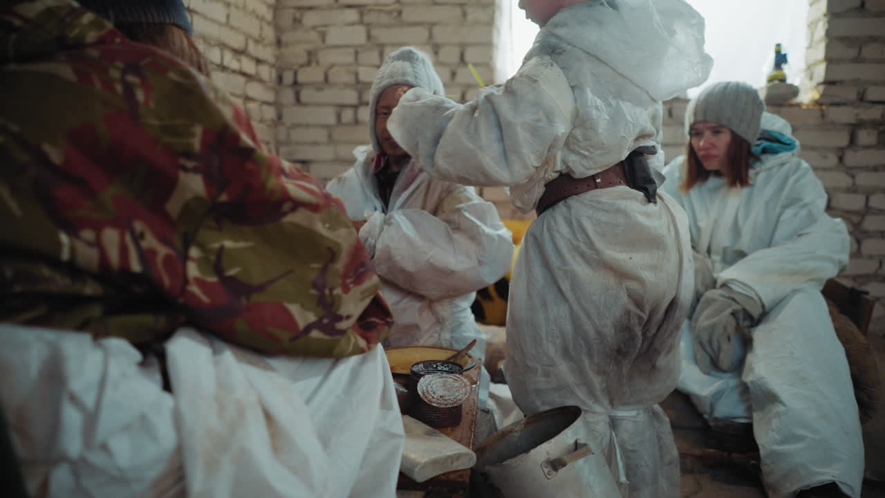 Yıpranmış koruyucu kıyafetler içindeki bir çocuk, harabe haldeki tuğla barınağın içinde hayatta kalanlar toplanıp kısıtlı kaynaklarla yemek hazırlarken otları bir tencereye koyuyor; savaş sonrası mücadele ve yoksulluk görülüyor