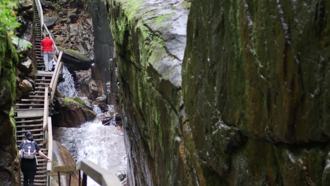폭포, 암벽, 나무 산책로를 오르는 사람들이 있는 협곡을 드러내기 위해 카메라가 움직일 때 돌담이 전경에 놓여 있습니다.