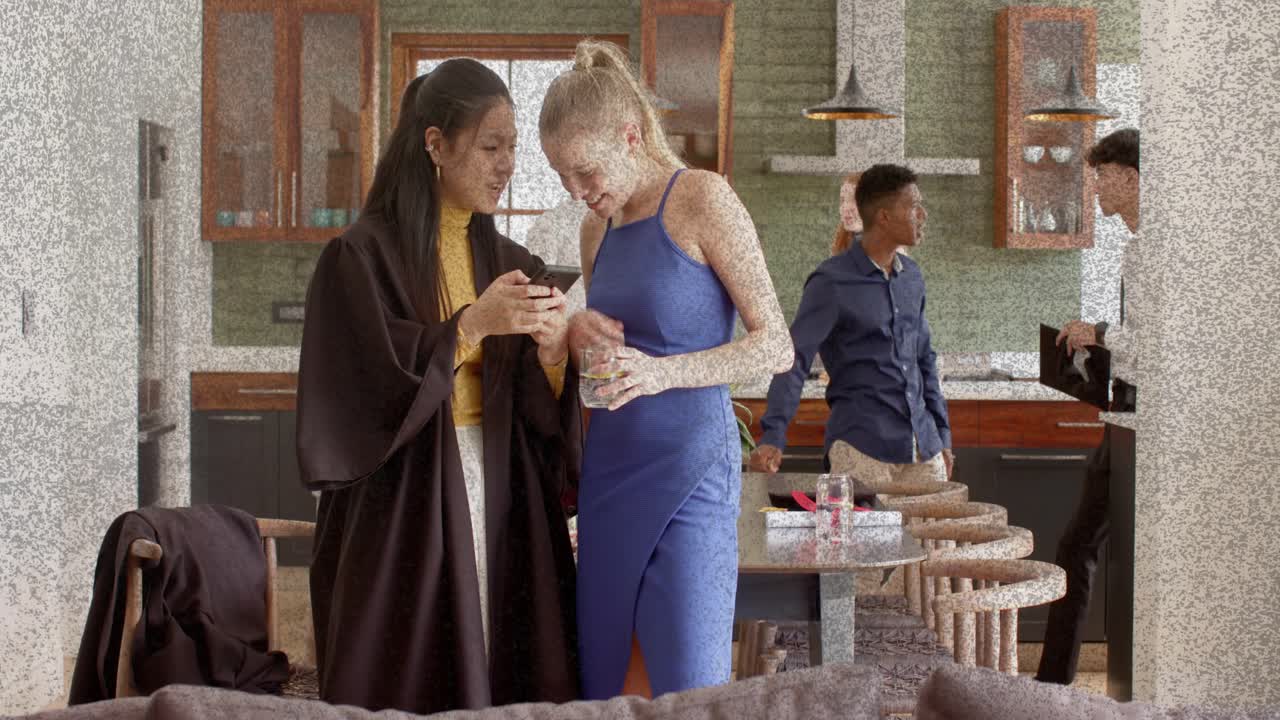 Left woman showing phone, right woman leaning, sharing clip, emojis popping near phone in kitchen