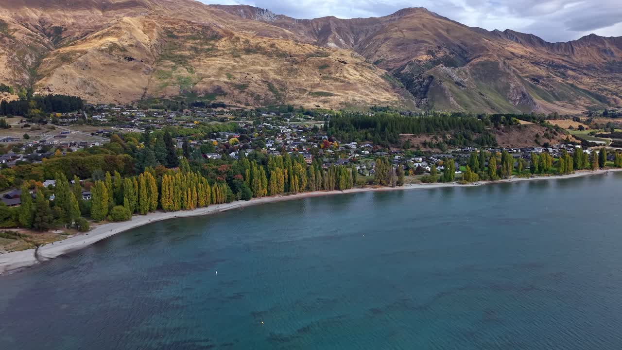 Aerial drone footage moving backward and ascending over Lake Wanaka in New Zealand, showing shoreline trees, town, and surrounding hills