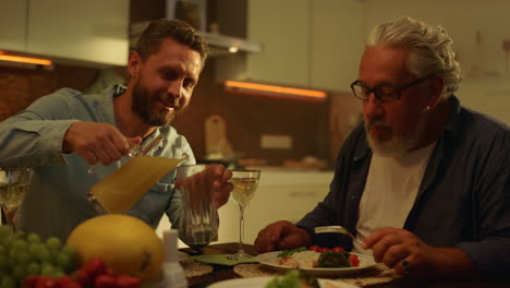 Senior-father-and-adult-son-eating-meal-at-table.-Man-pouring-juice-into-glass