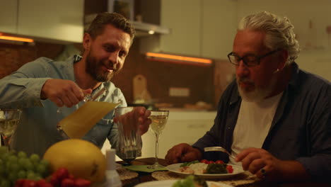 senior father and adult son eating meal at table. man pouring juice into glass