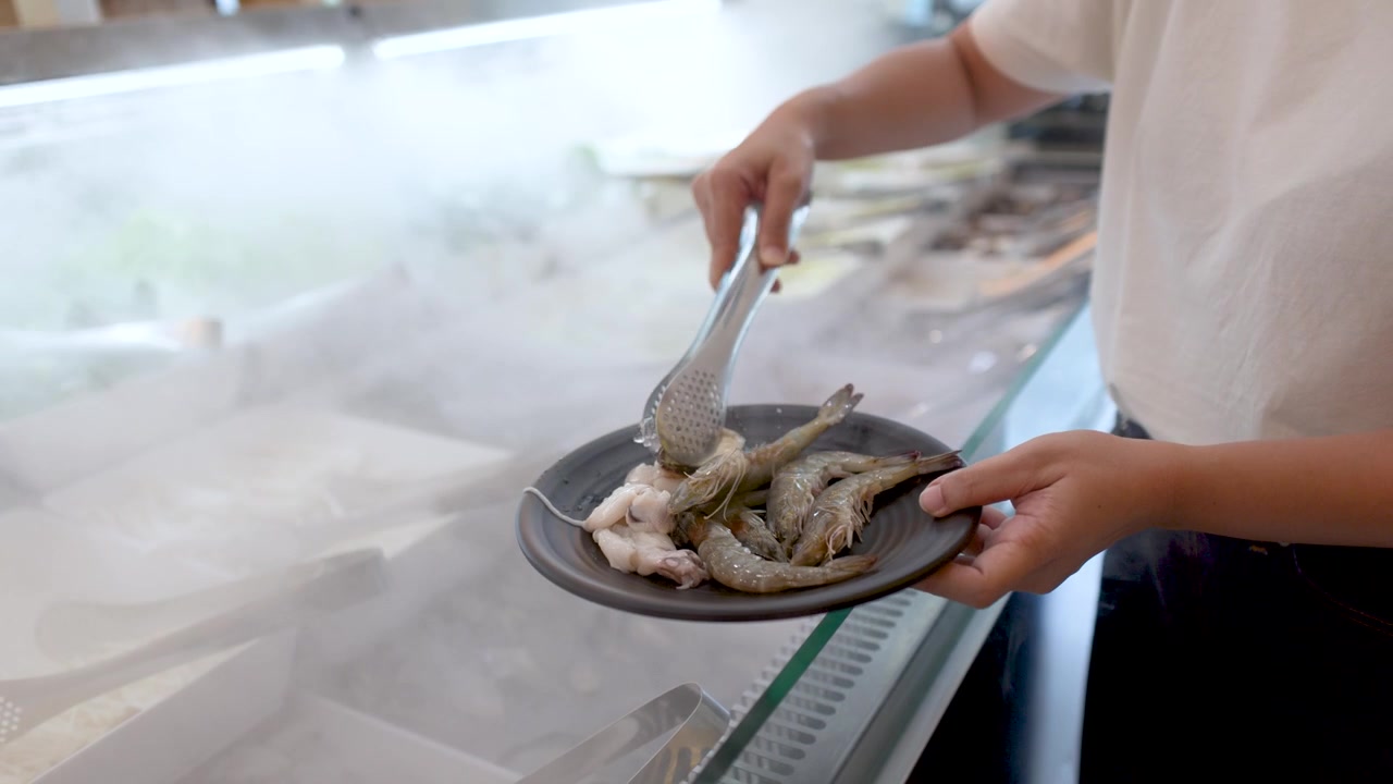 A person uses metal tongs to pick raw shrimp from a chilled buffet display counter