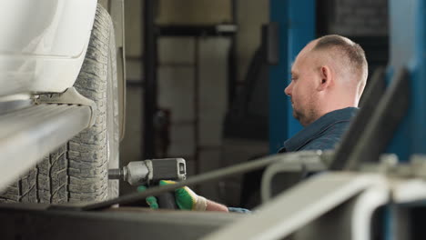 lower angle view of mechanic in blue uniform using pneumatic tool to tighten wheel bolts of lifted car in workshop, while partner approaches, inspects the work, and nods approvingly