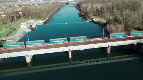 un largo tren de carga que transporta un contenedor de envío que conduce sobre un hermoso río en austria a través de un puente ferroviario