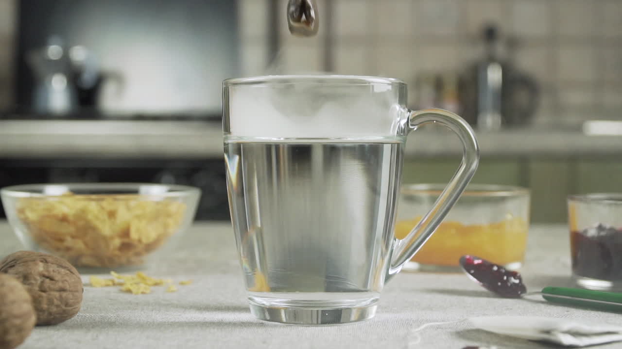 Una bolsita de té cae en una taza de agua caliente sobre una mesa preparada para el desayuno