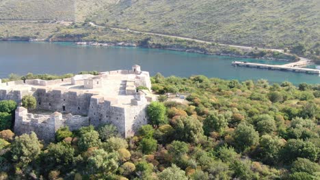 drone view in albania flying into a green island with a medieval fort on it on blue clear ocean water on a sunny day