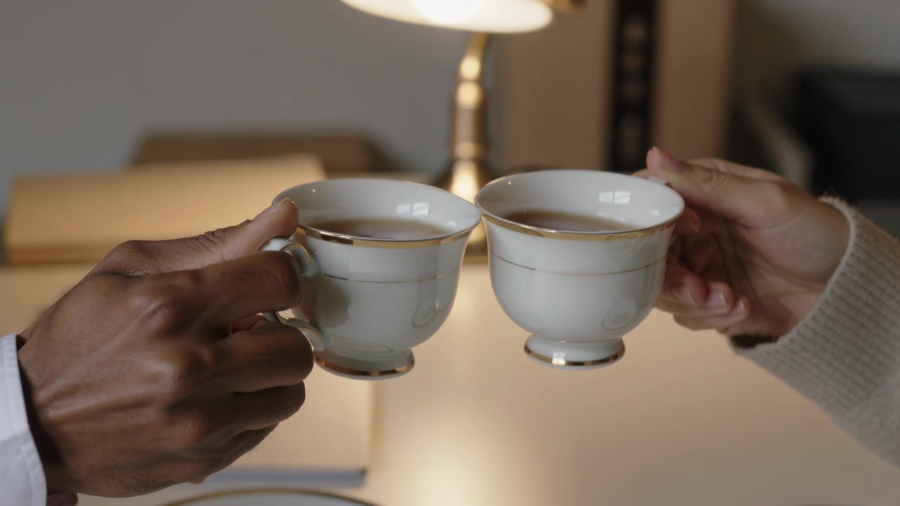 Entrant dans le cadre, un couple trinque avec des tasses à thé à bord doré sur une table, manchette de chemise, manche tricotée.