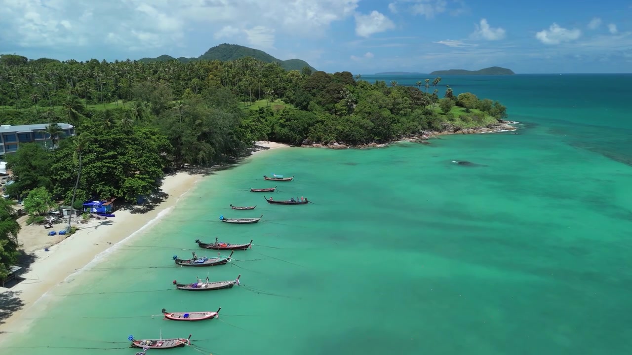 Sorvolo aereo con drone di Rawai Beach mentre una barca a coda lunga arriva vicino alla riva, con barche ancorate in acqua turchese, edifici costieri, colline verdi e isole vicine a Phuket