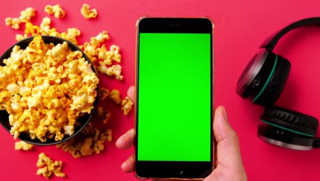 la mano del hombre sosteniendo un teléfono inteligente con pantalla táctil verde y palomitas de maíz en un tazón sobre fondo rojo.