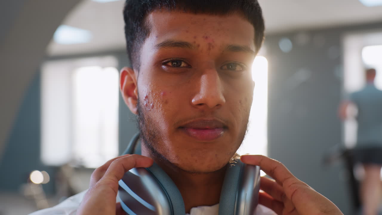 Close up of young man with visible acne and slight red eye removing headphones inside gym, with blurry background showing large windows, workout equipment, and person running on treadmill