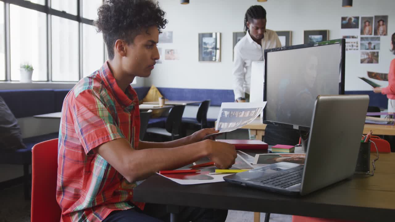 hombre de negocios biracial trabajando con una computadora portátil y sosteniendo una foto en la oficina