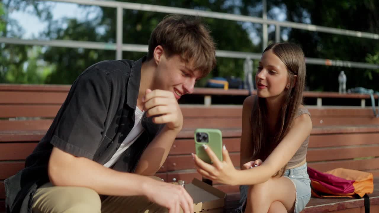 Two teenagers laugh together while looking at their phones outdoors
