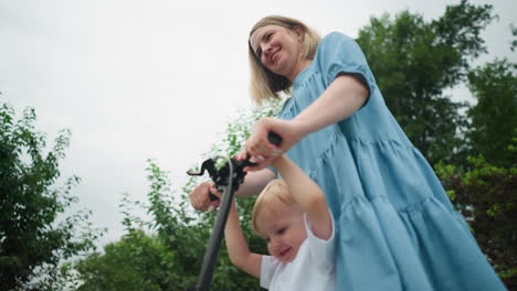 a mother and son ride a scooter together, both smiling warmly, close to a greenery background