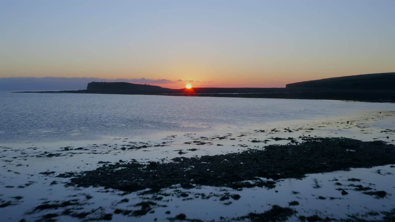 Aerial drone footage flying over the calm waters of Galway Bay at sunset, with warm colors and a silhouetted cliff.