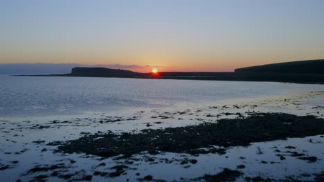 aerial drone footage flying over the calm waters of galway bay at sunset, with warm colors and a silhouetted cliff.