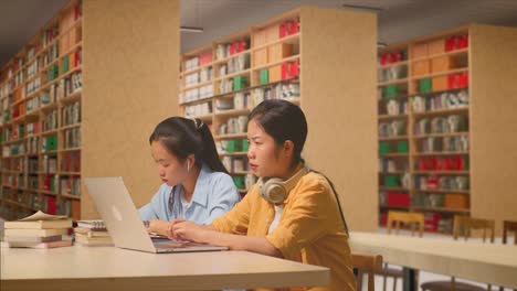 angry asian woman student with headphones typing on a laptop while sitting with her classmate studying on a table in the library