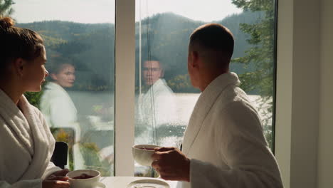 couple enjoys landscape looking in kitchen window. woman and man in white bathrobes drink herbal tea in lakeside glamping hotel. vacation at wild land