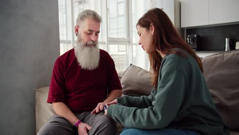 Un-Hombre-Mayor-De-Pelo-Gris-Y-Barba-Frondosa-Con-Una-Camiseta-Roja,-Junto-Con-Su-Hija-Morena-Adulta-Con-Una-Chaqueta-Verde,-Miden-Su-Pulso-Usando-Un-Dispositivo-Conectado-A-Su-Dedo-Mientras-Están-Sentados-En-El-Sofá-De-Un-Apartamento-Moderno.