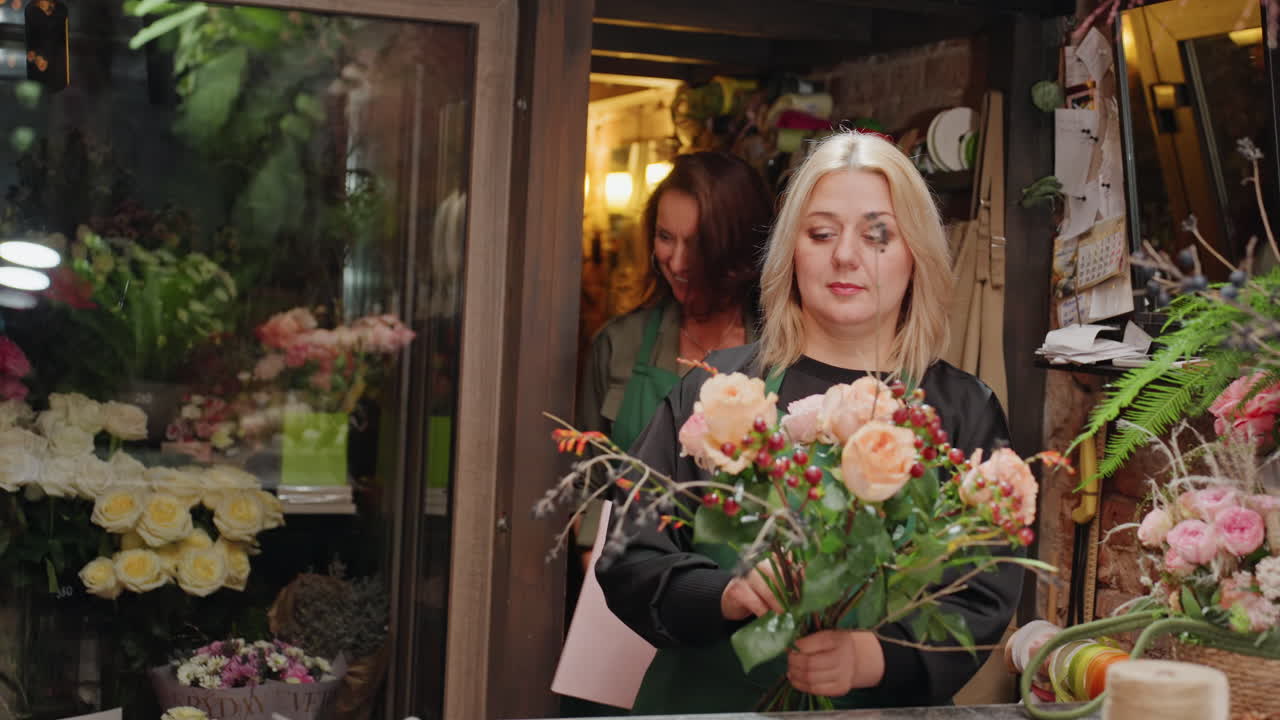 Fiorista bianca che dimostra la tecnica di legatura del bouquet, istruttore e apprendista che praticano metodi con filo e nastro, taglio attento degli steli, disposizione passo-passo sul bancone, risate e mani concentrate.