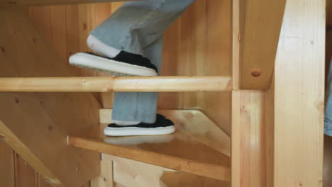 leg view of person in black slippers and denim pants walking down wooden staircase inside warm rustic cabin, soft indoor lighting emphasizes cozy atmosphere and movement through vertical space