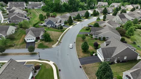 Toma-Aérea-De-Seguimiento-De-Un-Automóvil-Blanco-Que-Circula-Por-Una-Zona-Residencial-Moderna-Y-Noble-En-Verano