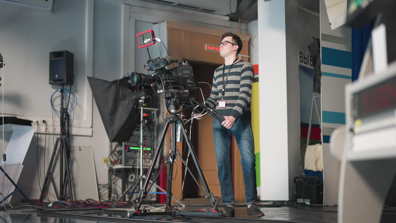 Camera operator walking in studio while carrying broadcast camera. Filming production setup in modern TV studio with various equipment and cables