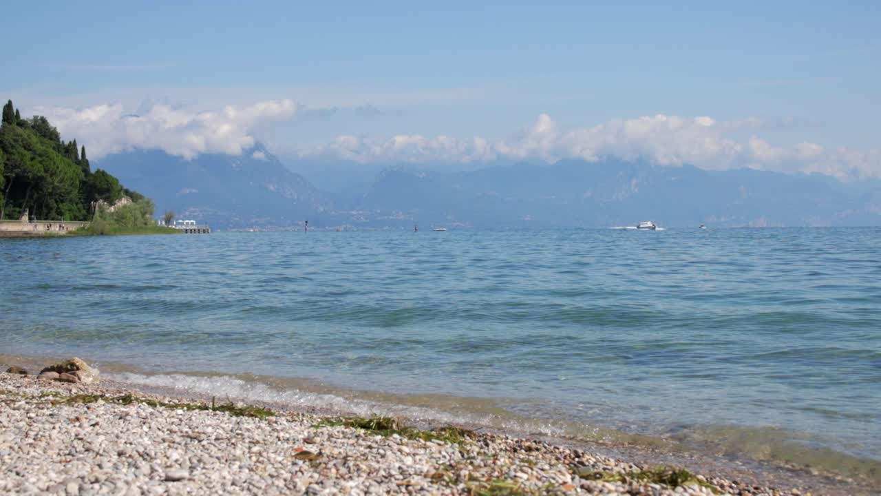 Small gentle waves crash on Lake Garda pebble rocky shores, clear green water of Italy