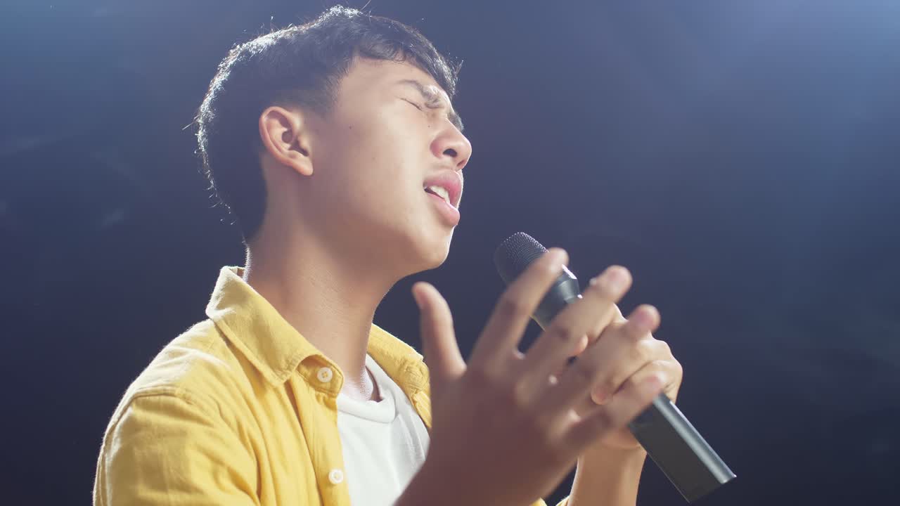 젊은 아시아 소년이 마이크를 들고  연기 검은 배경에서 노래하는 근접적인 측면 시각