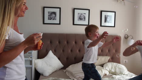 Caring-mother-plays-son-looking-at-soap-bubbles,-the-boy-is-happy-jumping-on-the-bed