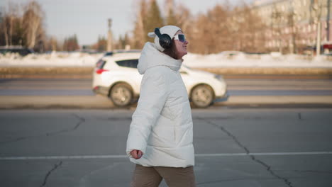 side view of young girl in white winter coat dancing on cracked pavement, arms relaxed in motion, surrounded by snowy urban scenery, vehicles in motion, and sunlight casting soft shadows